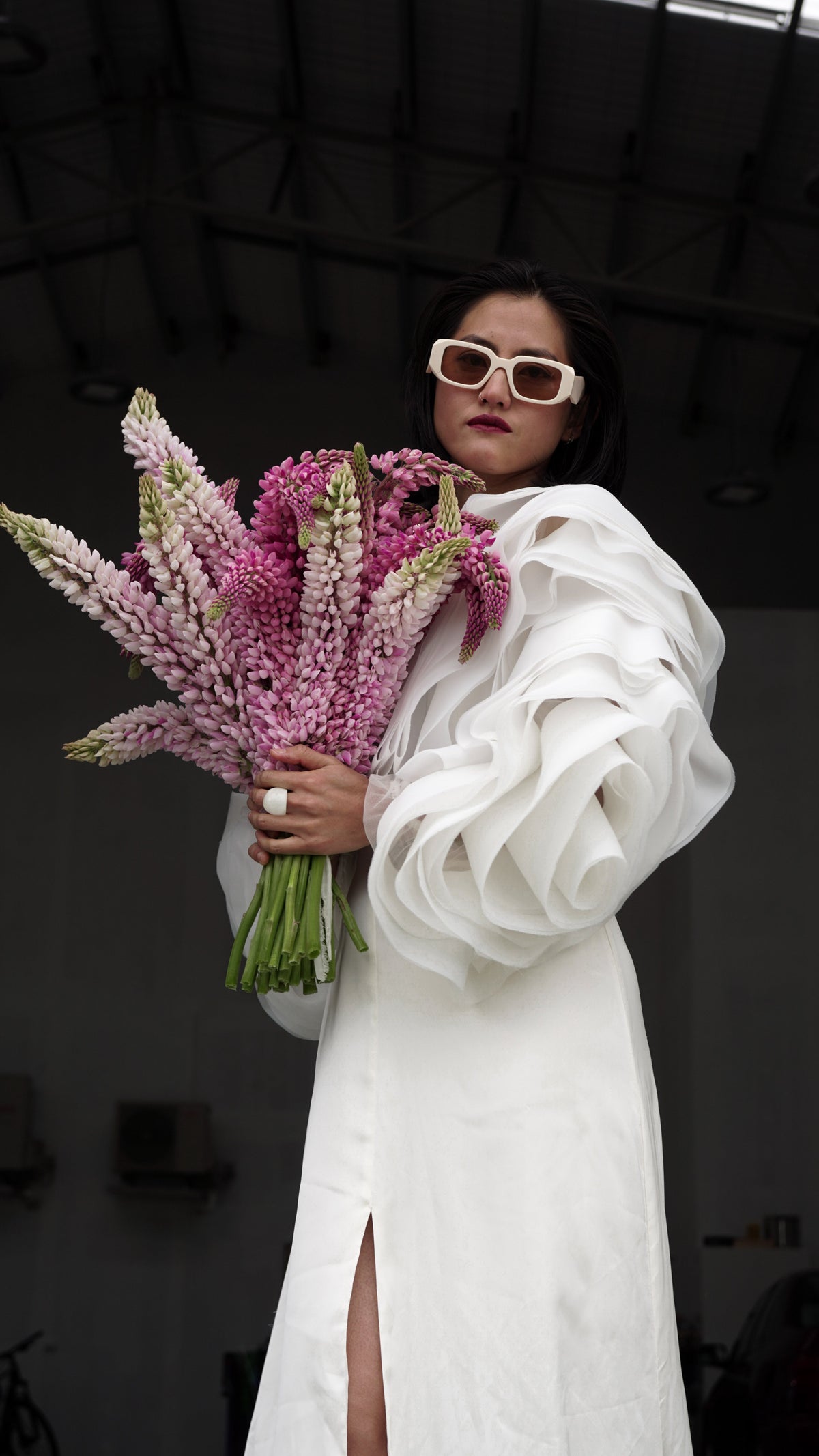 Bridal jacket with layered petal sleeves over a white slit dress, model holding pink lupines and wearing sunglasses, My Darling Hurricane