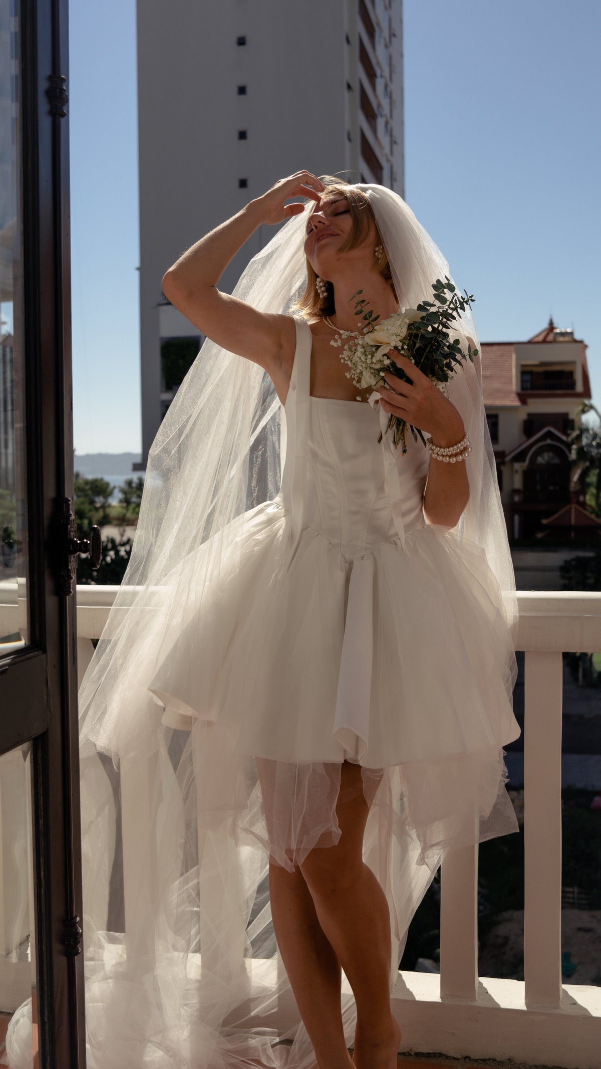 Bride shading her eyes on a sunny balcony, plain cathedral wedding veil framing a corset mini dress and bouquet, Niagara Veil