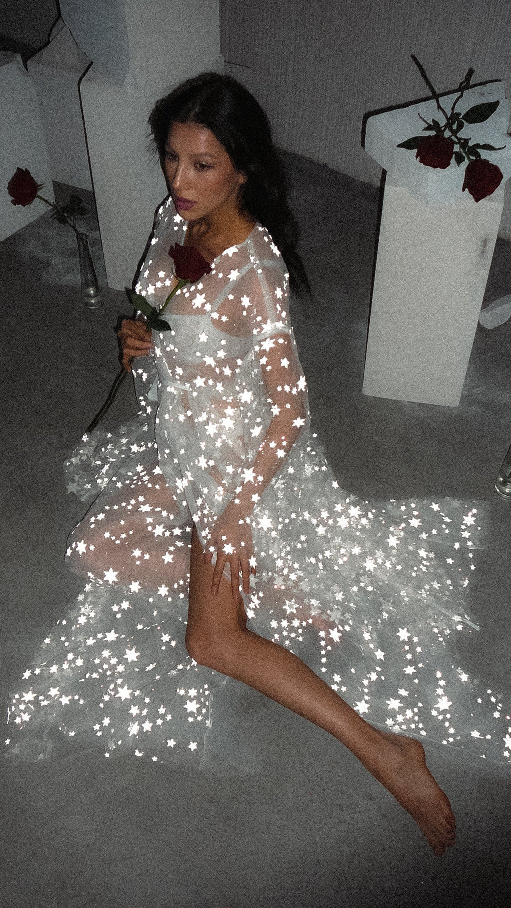 Model holding a red rose to her lips, sitting in a star tulle bridal robe on a concrete floor, Orion Robe