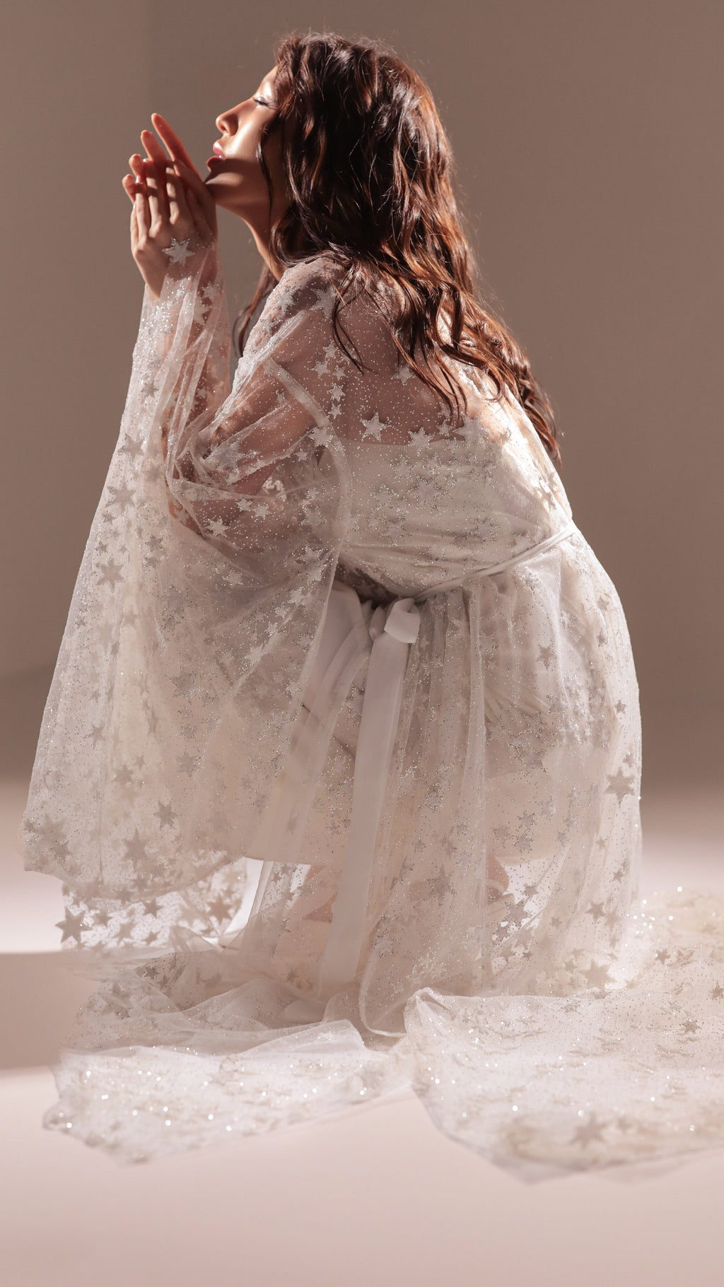 Model kneeling in profile in a star tulle bridal robe with a ribbon tie, hands raised to her face, Orion Robe