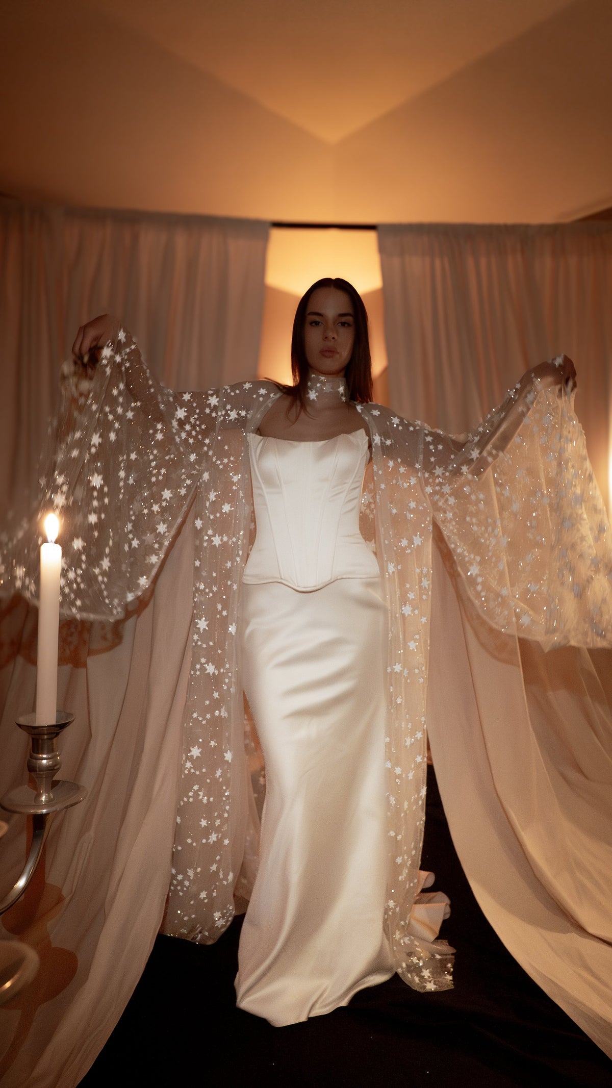 Bride in a satin corset gown with a sheer star bridal robe held open like wings, candles and drapes behind, Orion Robe