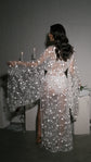 Back view of a sheer star bridal robe with bell sleeves and a ribbon belt, model facing lit candles, Orion Robe