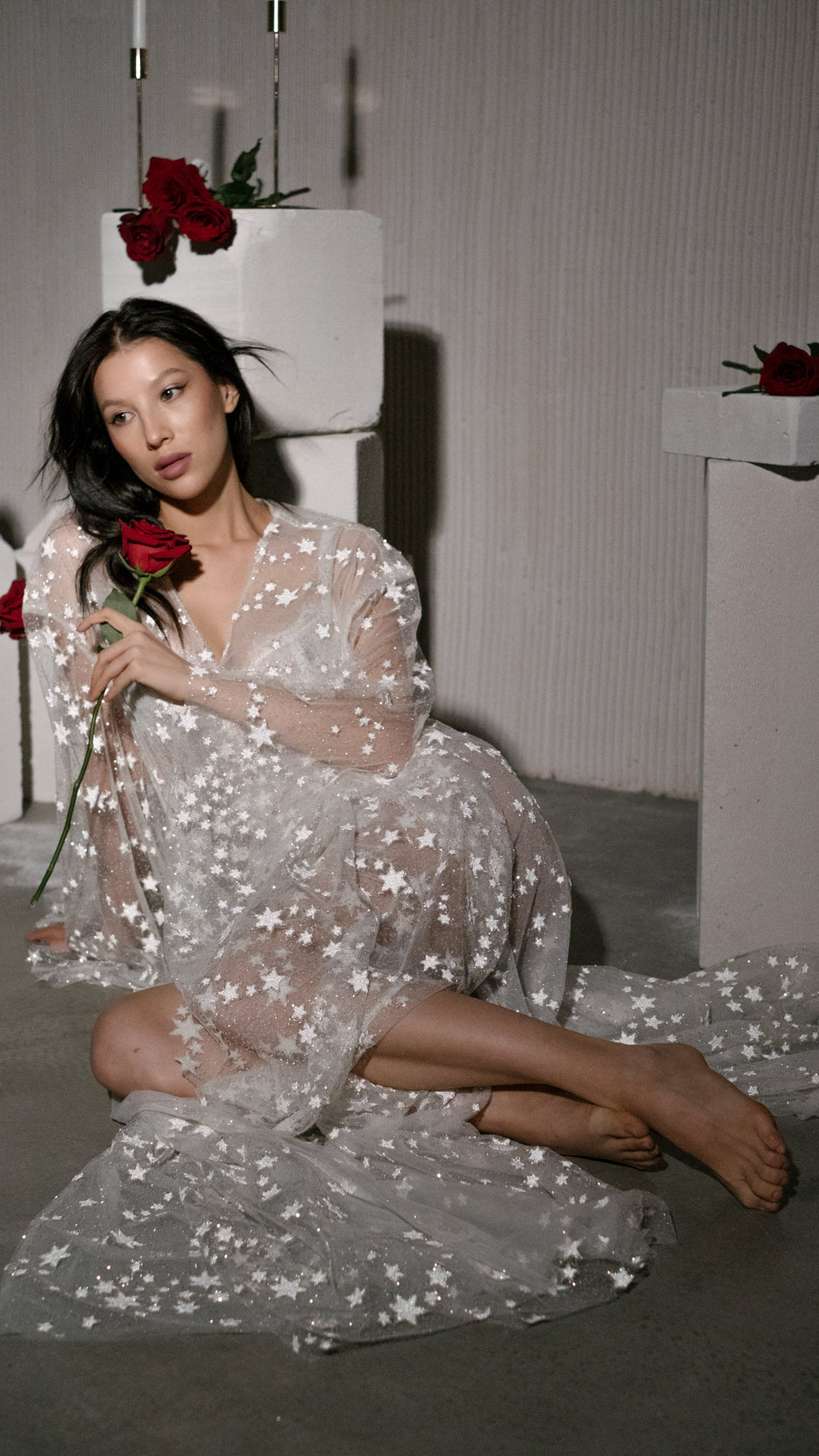 Model sitting cross-legged in a starry bridal robe with a red rose, roses on white plinths behind, Orion Robe