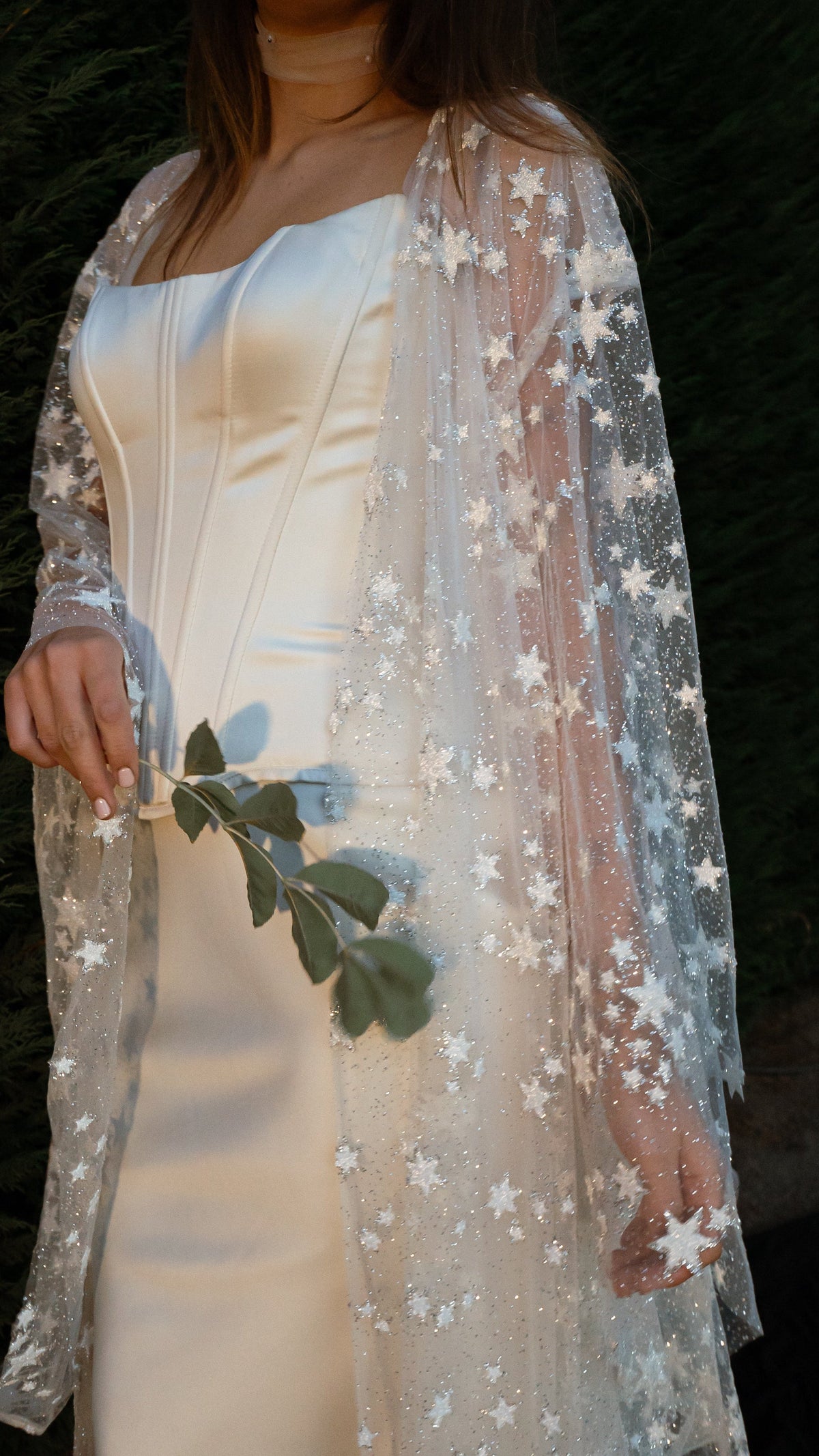 Star tulle bridal robe over a satin corset gown, bride holding a leafy stem, cropped torso shot at night, Orion Robe