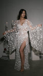 Model in a star tulle bridal robe with a high slit, one sleeve held out, candles and red roses on white plinths, Orion Robe