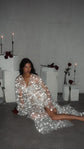 Model reclining on the floor in a sheer starry bridal robe, roses and candles on white blocks behind her, Orion Robe