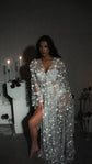 Model seated on a white plinth in a sheer star bridal robe, bare leg through the slit, candles and roses, Orion Robe