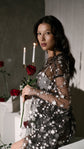 Model seated with a red rose in a sheer black star bridal robe, candles and roses on white blocks, Orion Robe Black
