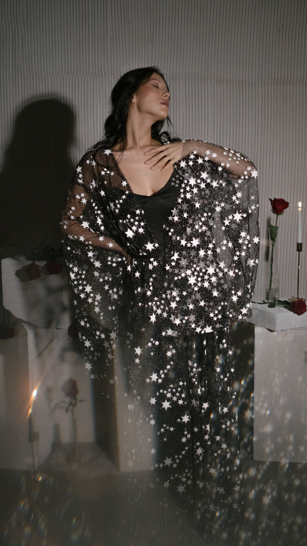 Model with eyes closed and hand at her chest in a black star bridal robe over a black slip, blurred foreground, Orion Robe Black