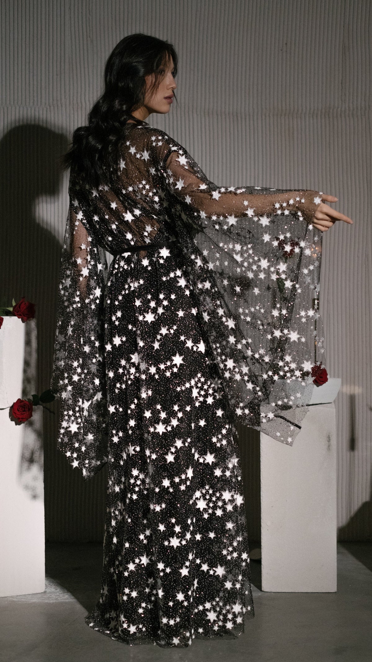 Back view of a black star bridal robe with sweeping bell sleeves, model reaching toward a white plinth, Orion Robe Black