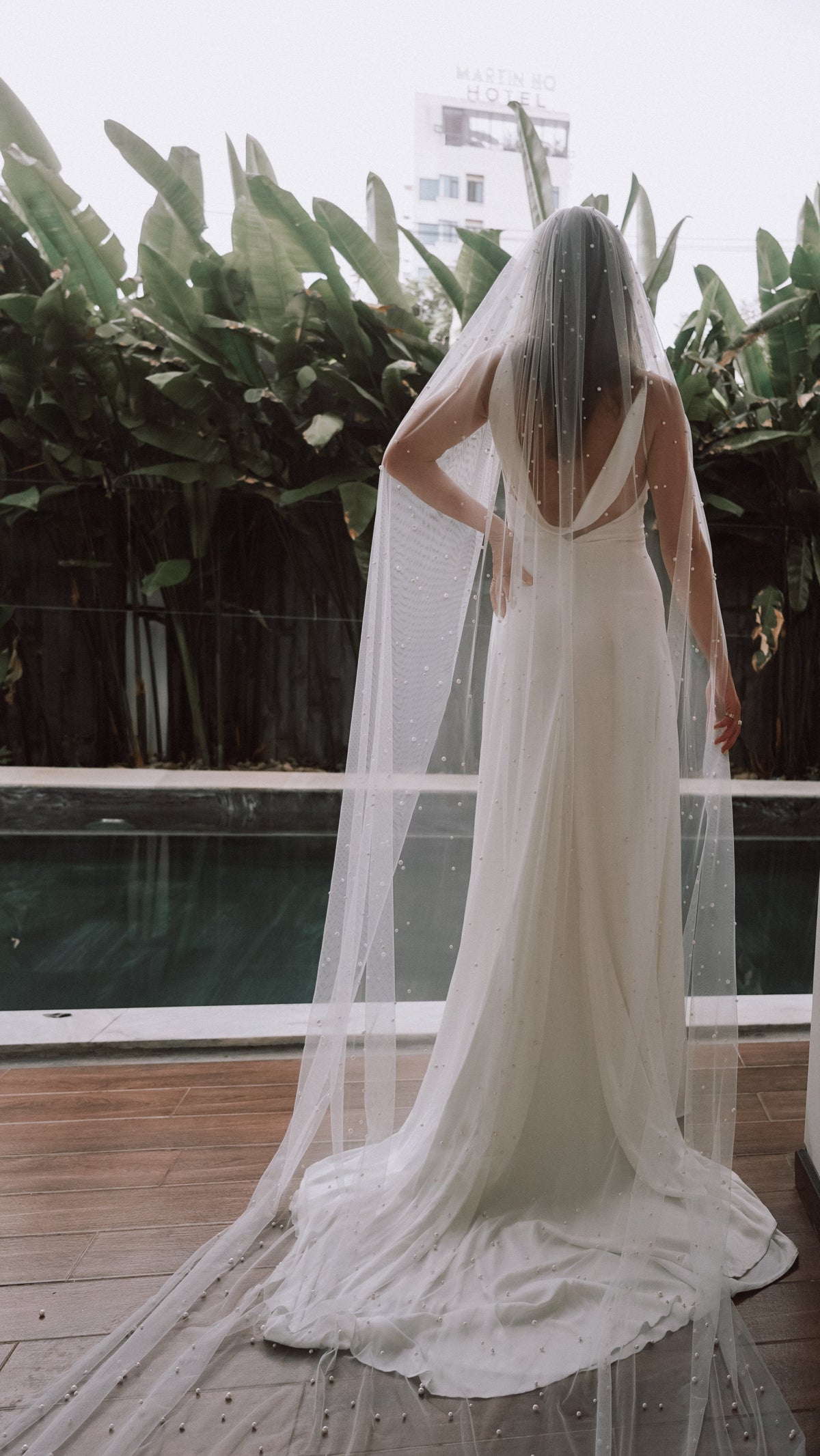 Cathedral wedding veil scattered with pearls trailing over a poolside deck, back view of a bride in a cowl-back gown, Pearl Cascade Veil by Ephemeris