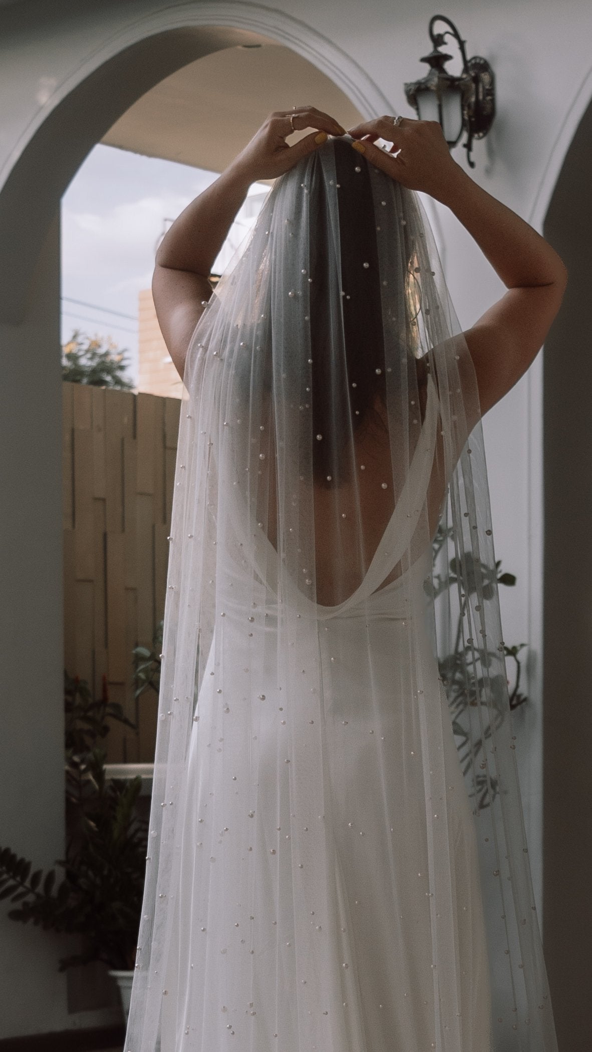 Bride adjusting a pearl cathedral wedding veil at the crown, back view under a white arch, Pearl Cascade Veil