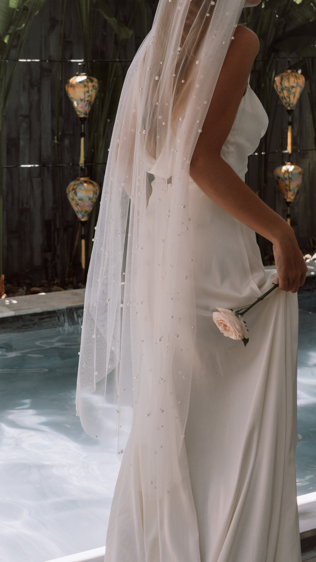 Fingertip wedding veil with scattered pearls over a sleek slip gown, bride by a pool holding a pale rose, Pearl Whispers Veil by Ephemeris