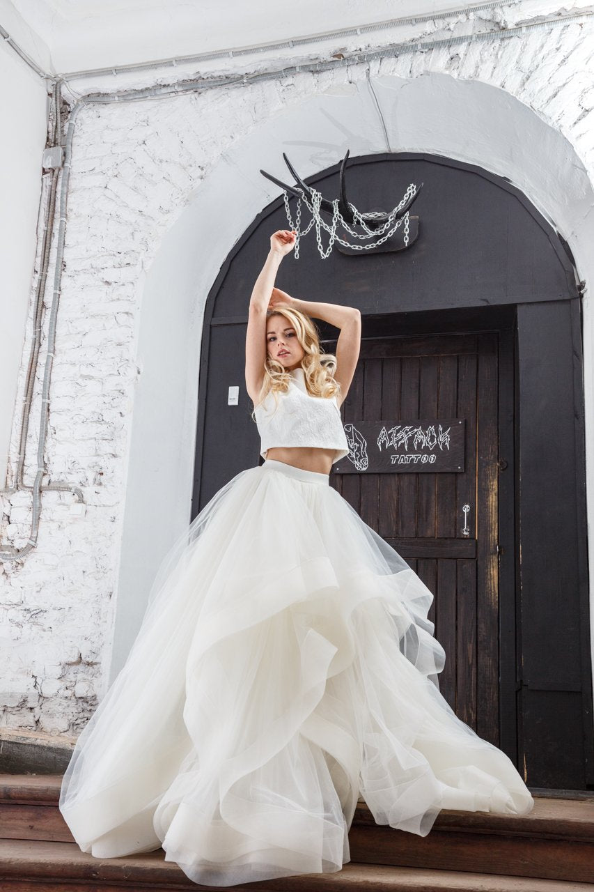 Bride with arms raised in a crop top and tiered tulle skirt, ivory two-piece wedding dress, white brick arch, Ready or Not Ivory