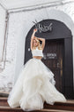 Bride with arms raised in a crop top and tiered tulle skirt, ivory two-piece wedding dress, white brick arch, Ready or Not Ivory