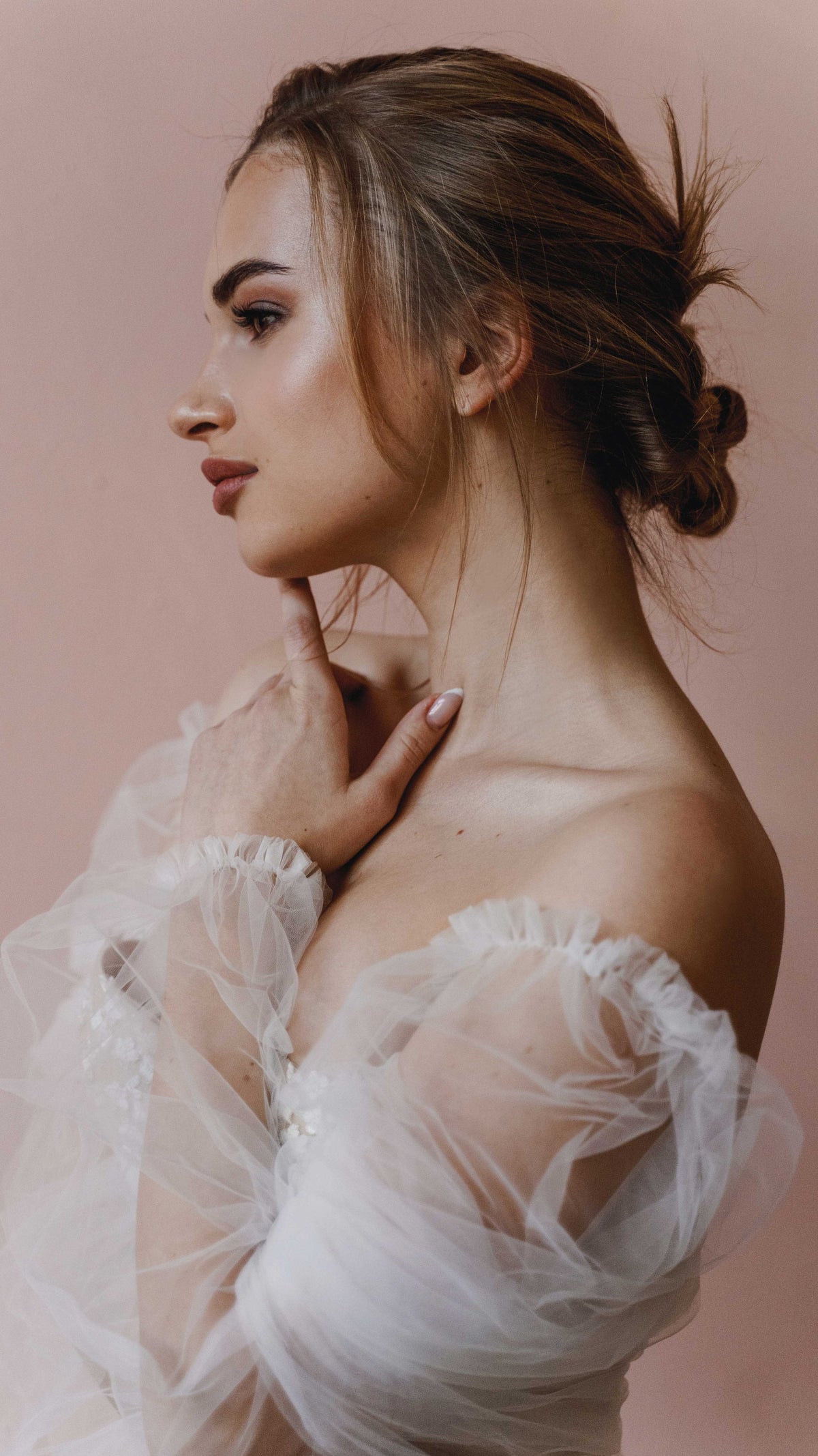 Side profile of a bride touching her neck, tulle puff sleeve of an off-shoulder wedding dress, pink backdrop, Savanna