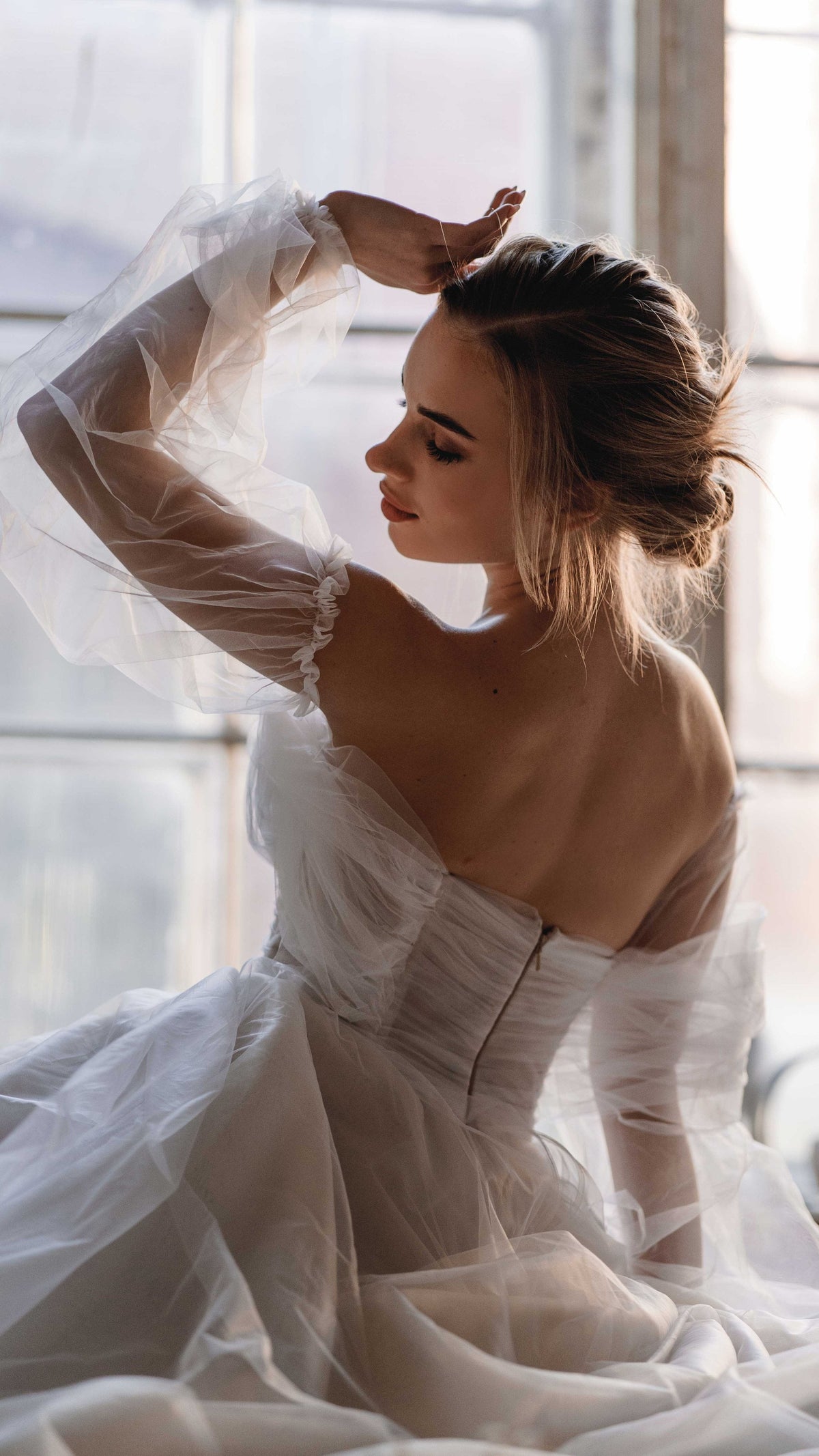 Back view of an off-shoulder wedding dress with a boned corset and sheer puff sleeves, bride by a bright window, Savanna