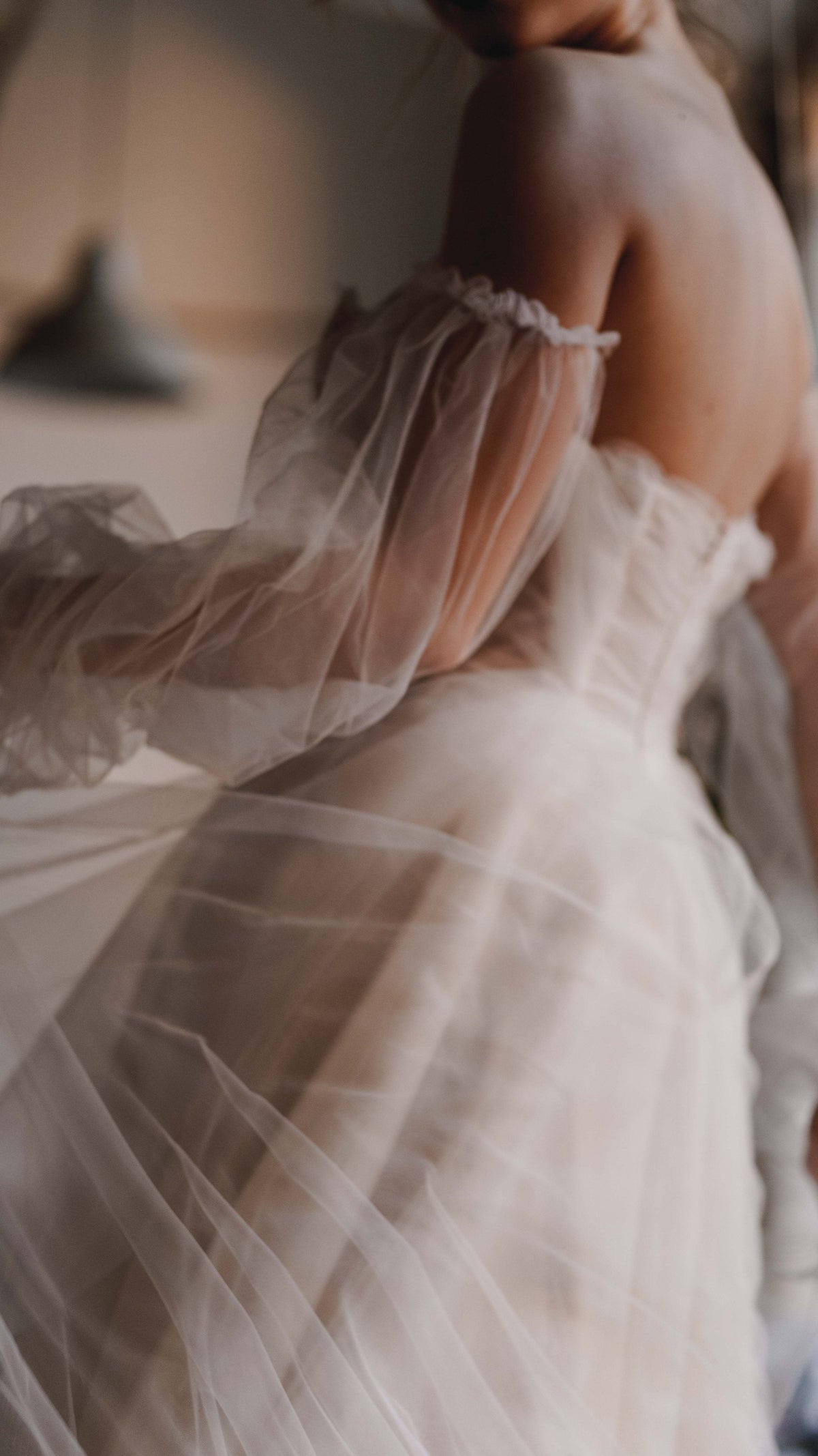 Soft-focus back detail of a tulle wedding dress with detachable puff sleeves slipping off the shoulder, Savanna