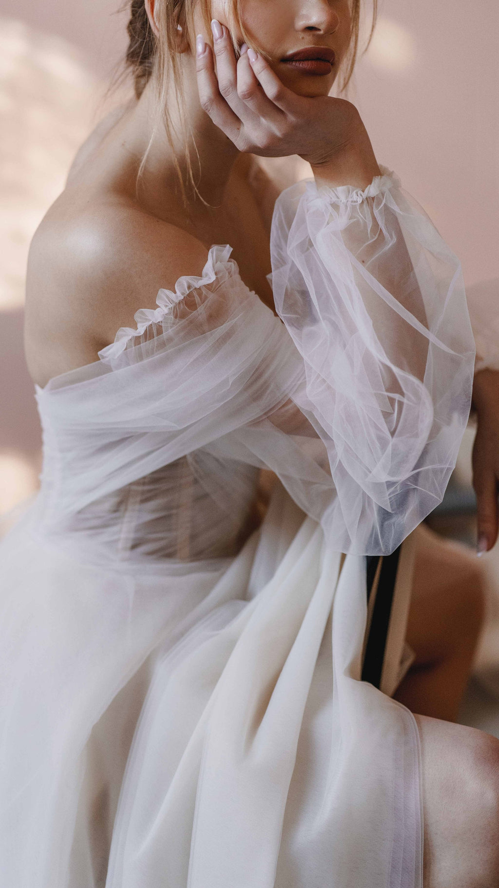 Close crop of a seated bride in an off-shoulder tulle wedding dress, ruffled puff sleeve and hand at her cheek, Savanna