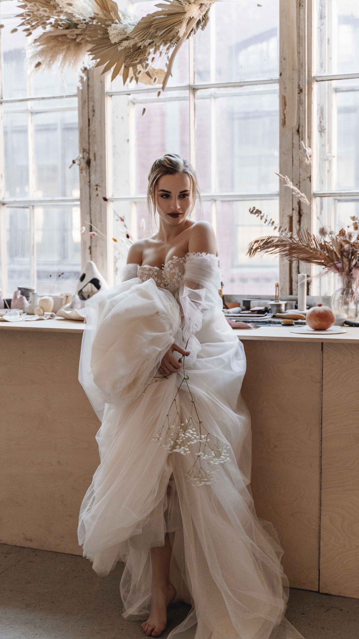 Bride by a sunny window lifting the tulle skirt of an off-shoulder ball gown wedding dress, dried pampas above, Savanna