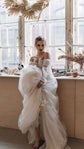 Bride by a sunny window lifting the tulle skirt of an off-shoulder ball gown wedding dress, dried pampas above, Savanna