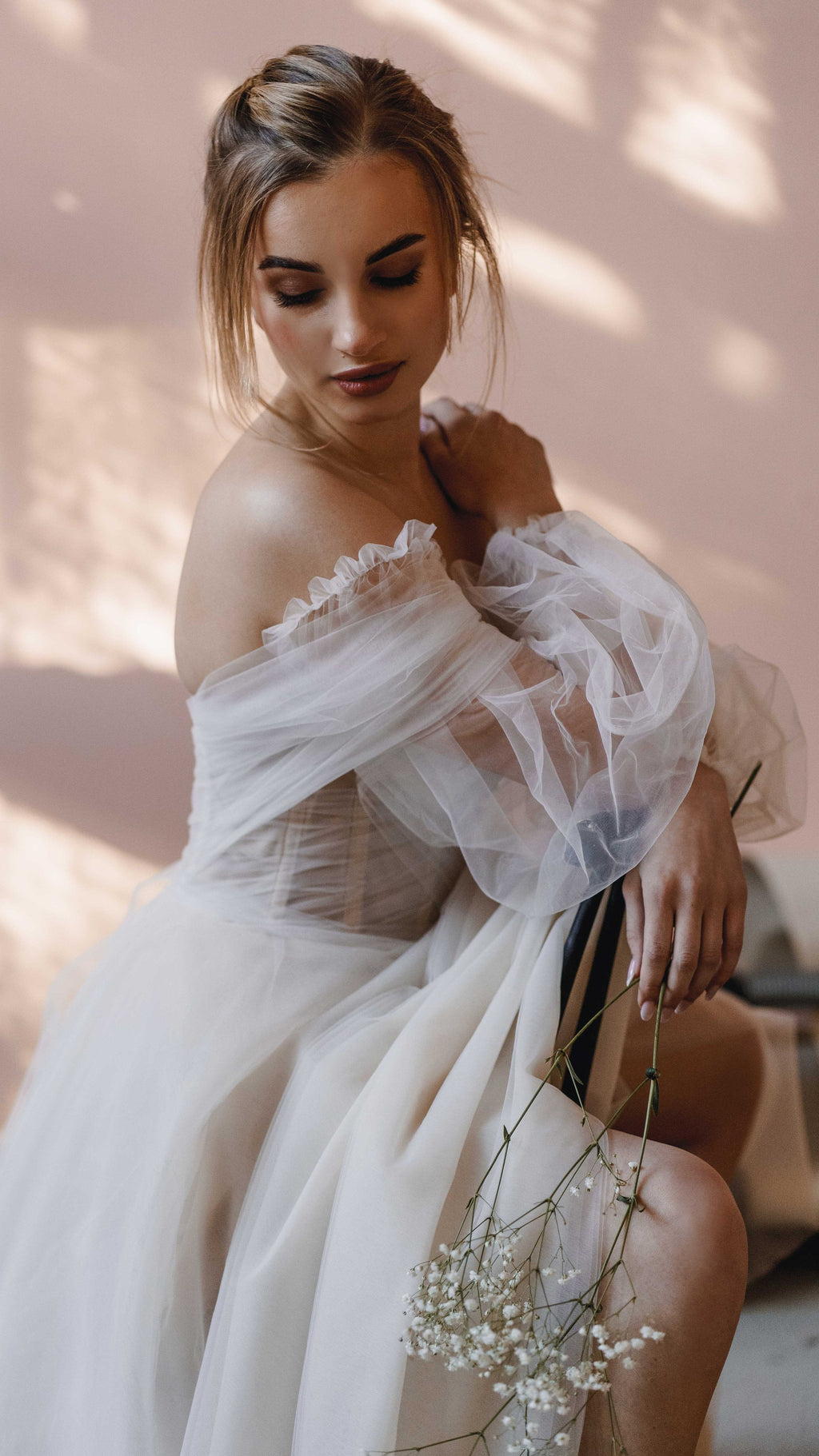 Seated bride holding baby's breath, off-shoulder wedding dress with sheer tulle puff sleeves, pink wall, Savanna