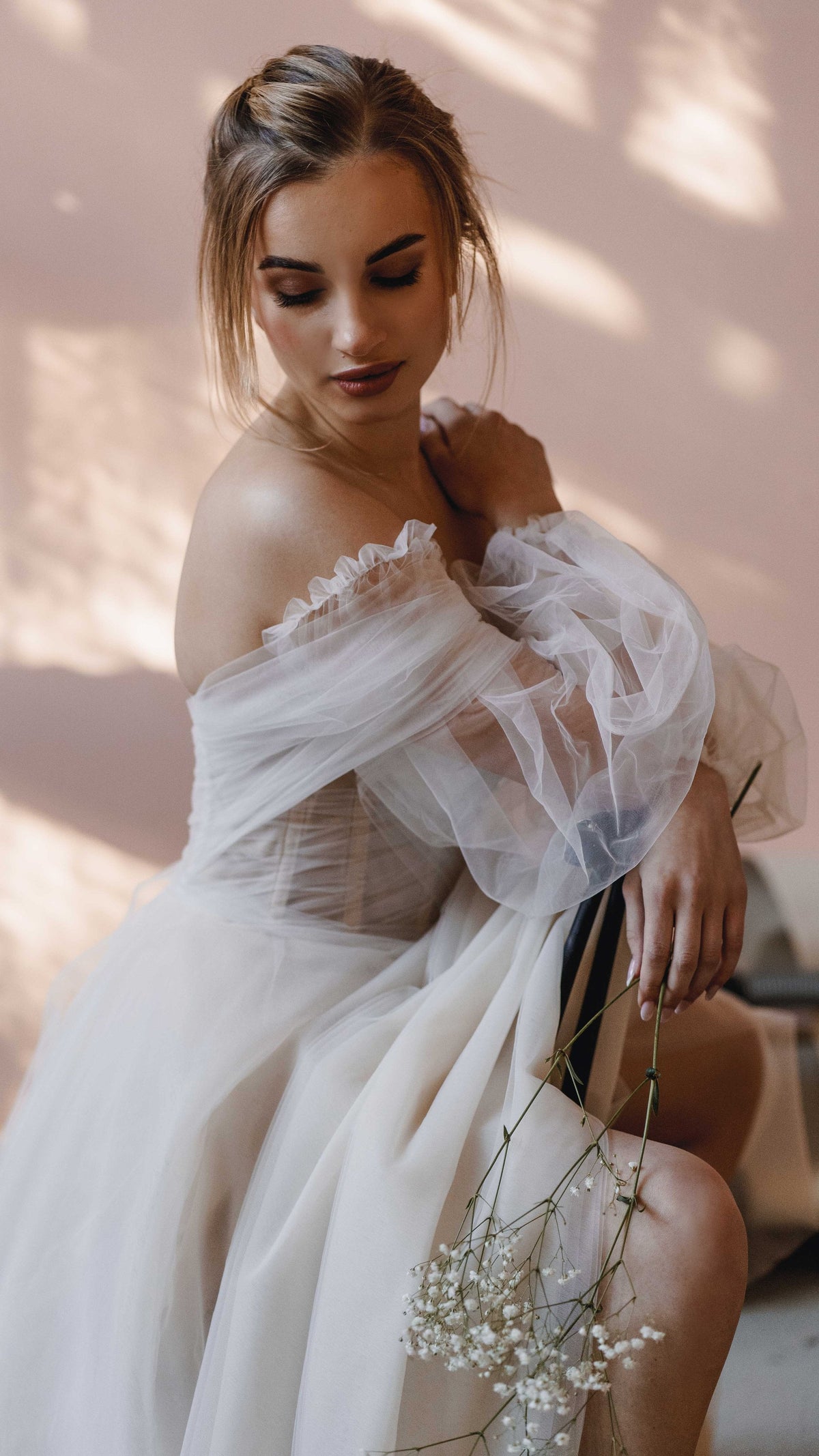 Seated bride holding baby's breath, off-shoulder wedding dress with sheer tulle puff sleeves, pink wall, Savanna