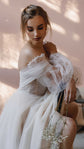 Seated bride holding baby's breath, off-shoulder wedding dress with sheer tulle puff sleeves, pink wall, Savanna