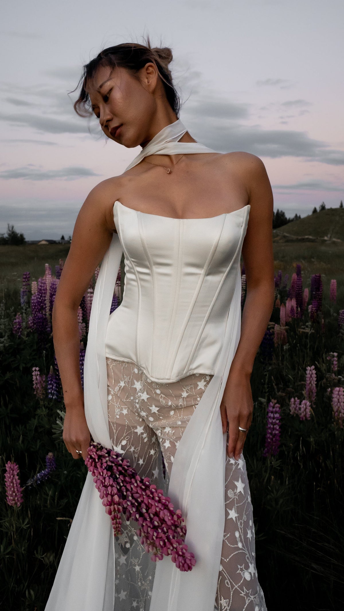 Strapless satin bridal corset with a basque waist, worn with star lace trousers and a chiffon scarf in a lupin field at dusk, Solana Corset by Ephemeris