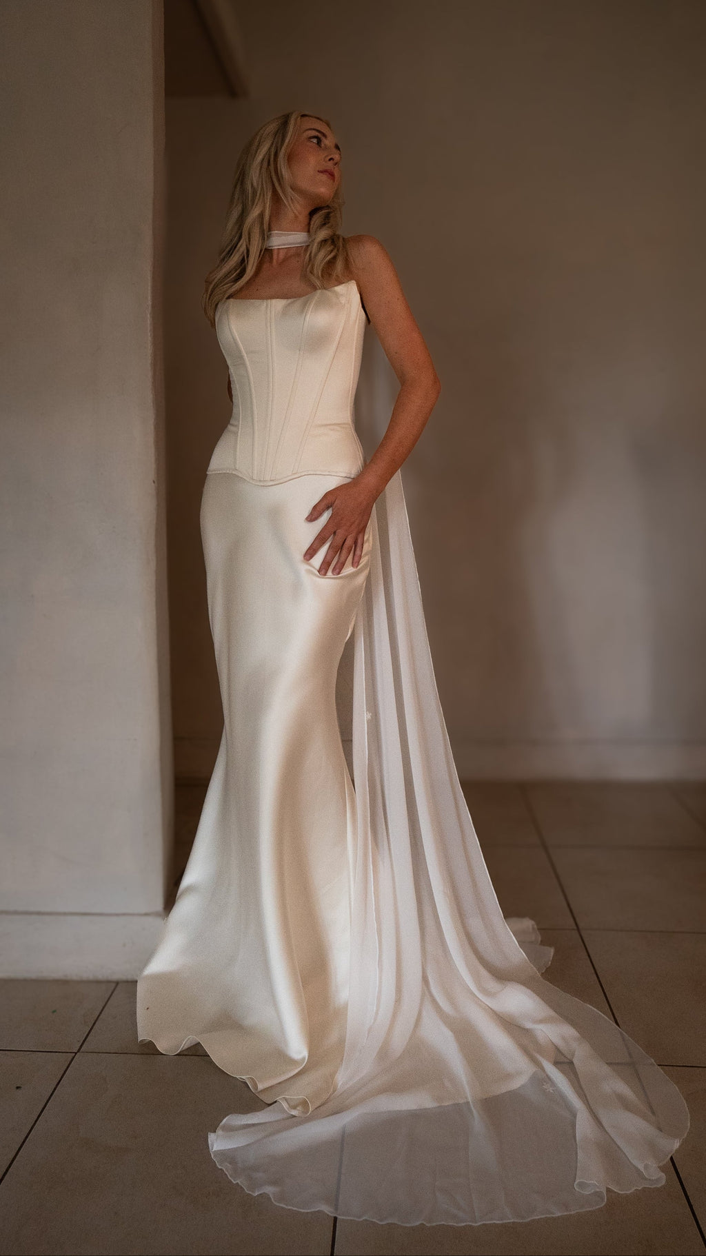 Bride gazing upward in a strapless bridal corset with a chiffon scarf flowing into the satin train, warm room, Solana Corset