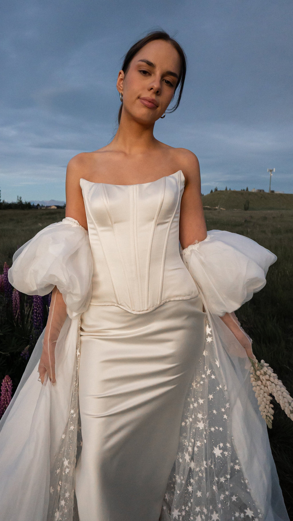 Low-angle portrait of a bride in a strapless bridal corset with puff sleeves and star tulle train against an open sky, Solana Corset