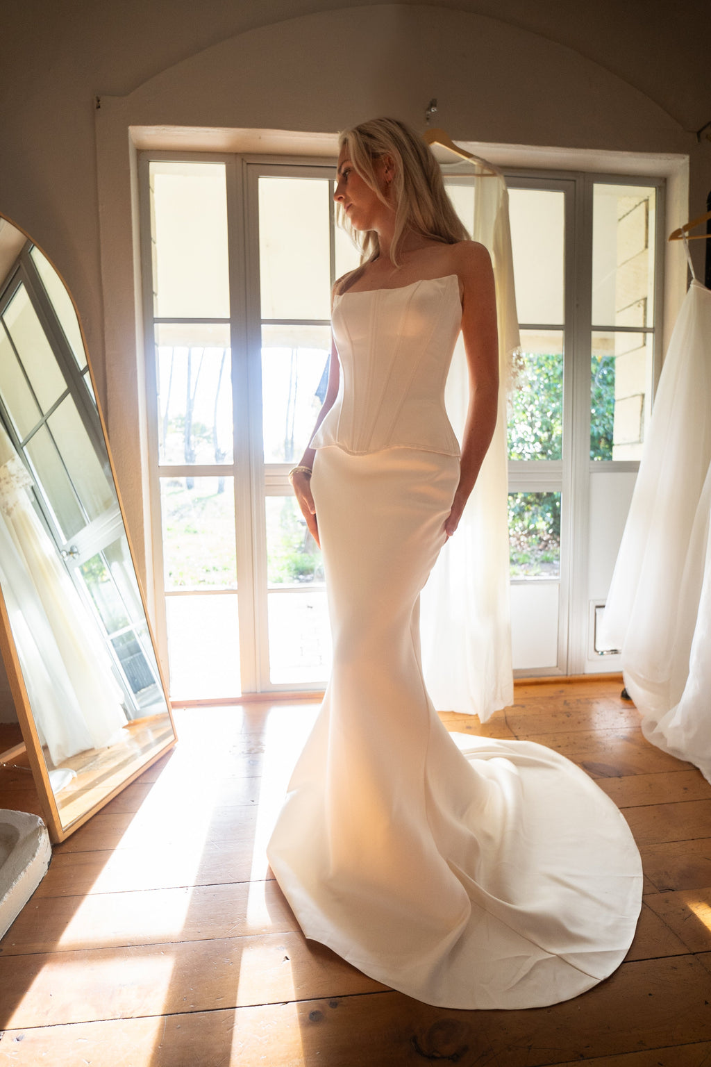 Corset and skirt bridal separates in satin, bride by sunlit French doors with a long train spread on floorboards, Solana Ensemble