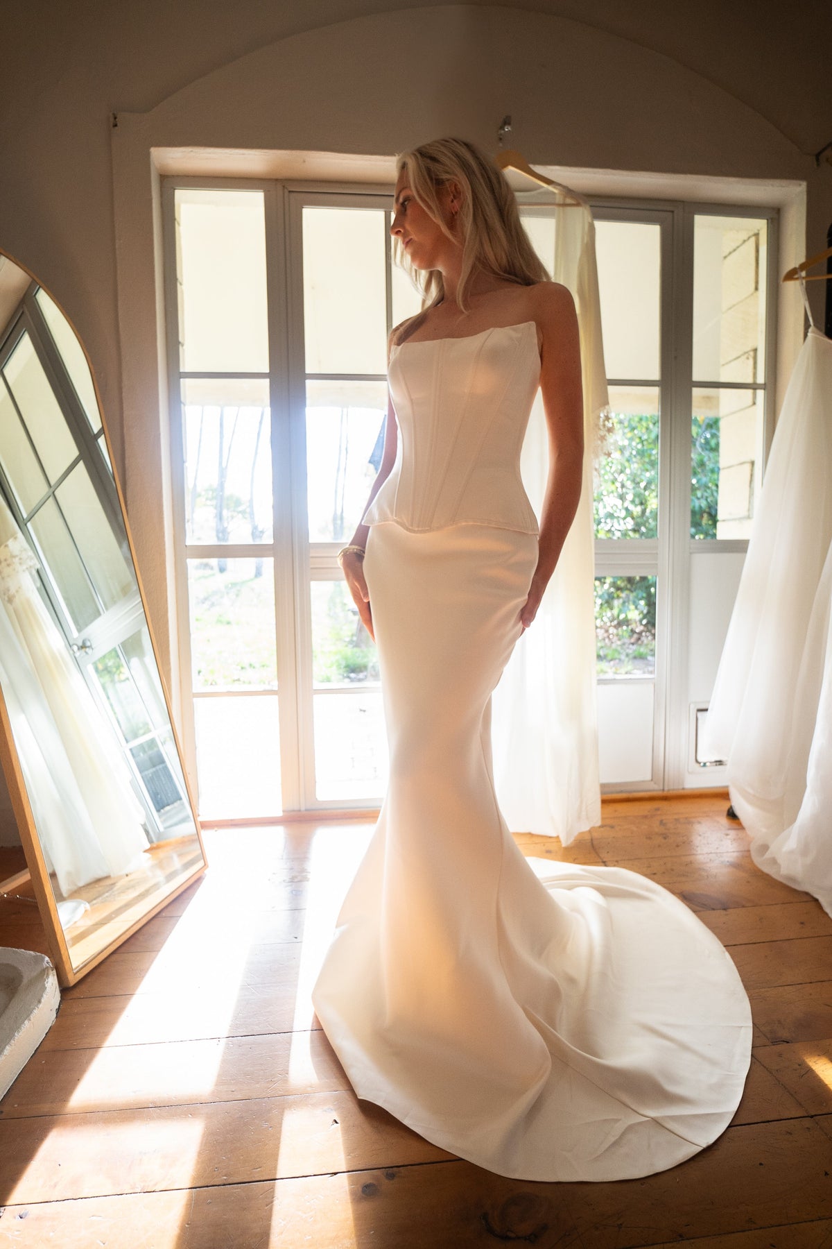 Corset and skirt bridal separates in satin, bride by sunlit French doors with a long train spread on floorboards, Solana Ensemble
