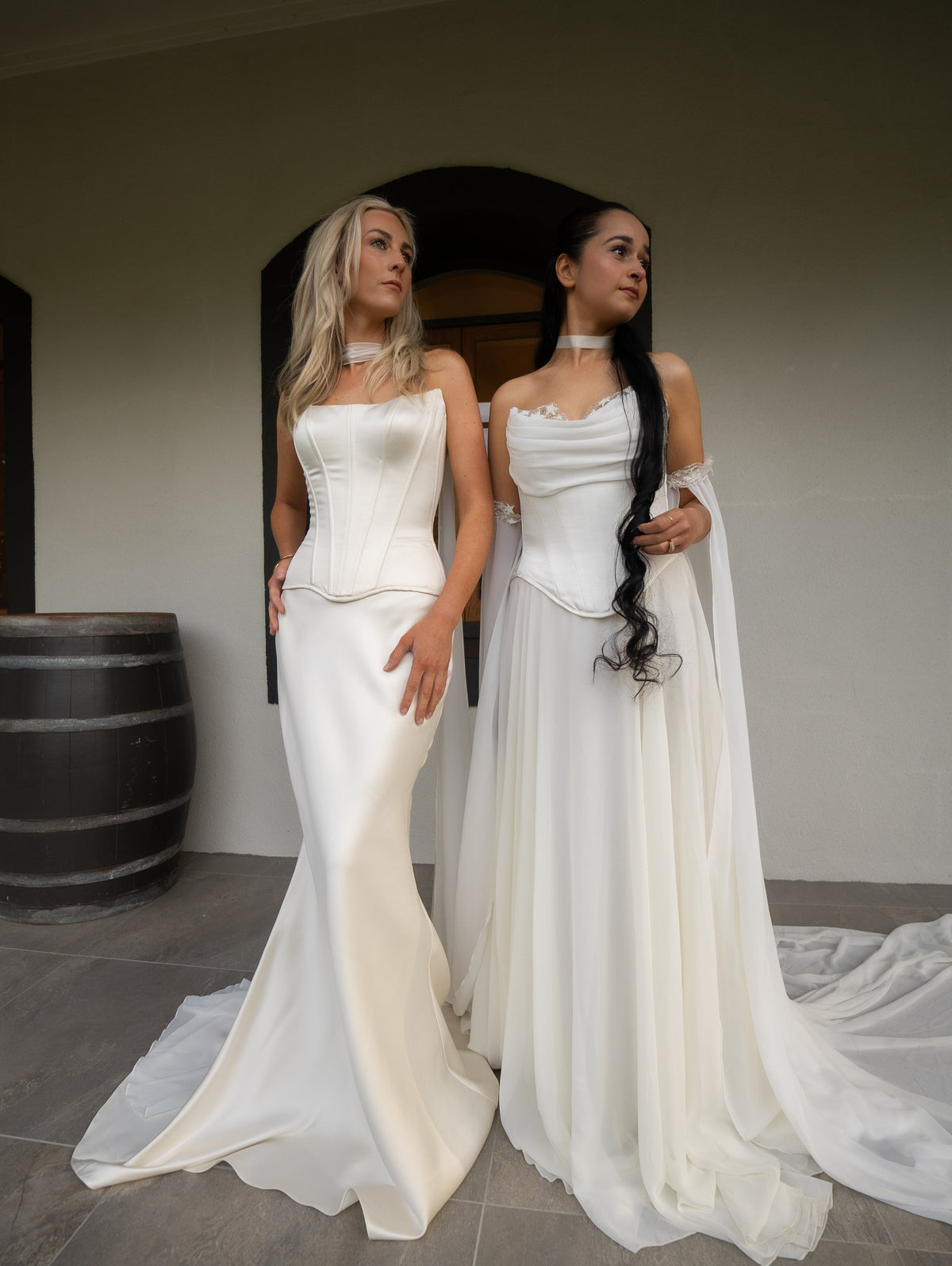 Two brides in a winery hall, one in satin corset and skirt bridal separates, the other in an ivory gown, Solana Ensemble