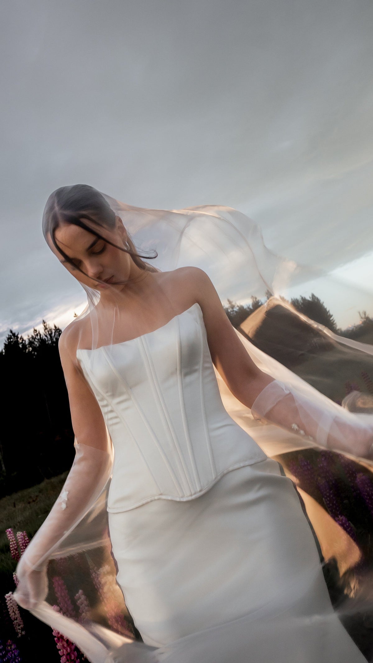 Bride in a satin bridal skirt and corset, a sheer tulle veil swept across her face by the wind, cloudy sky, Solana Skirt
