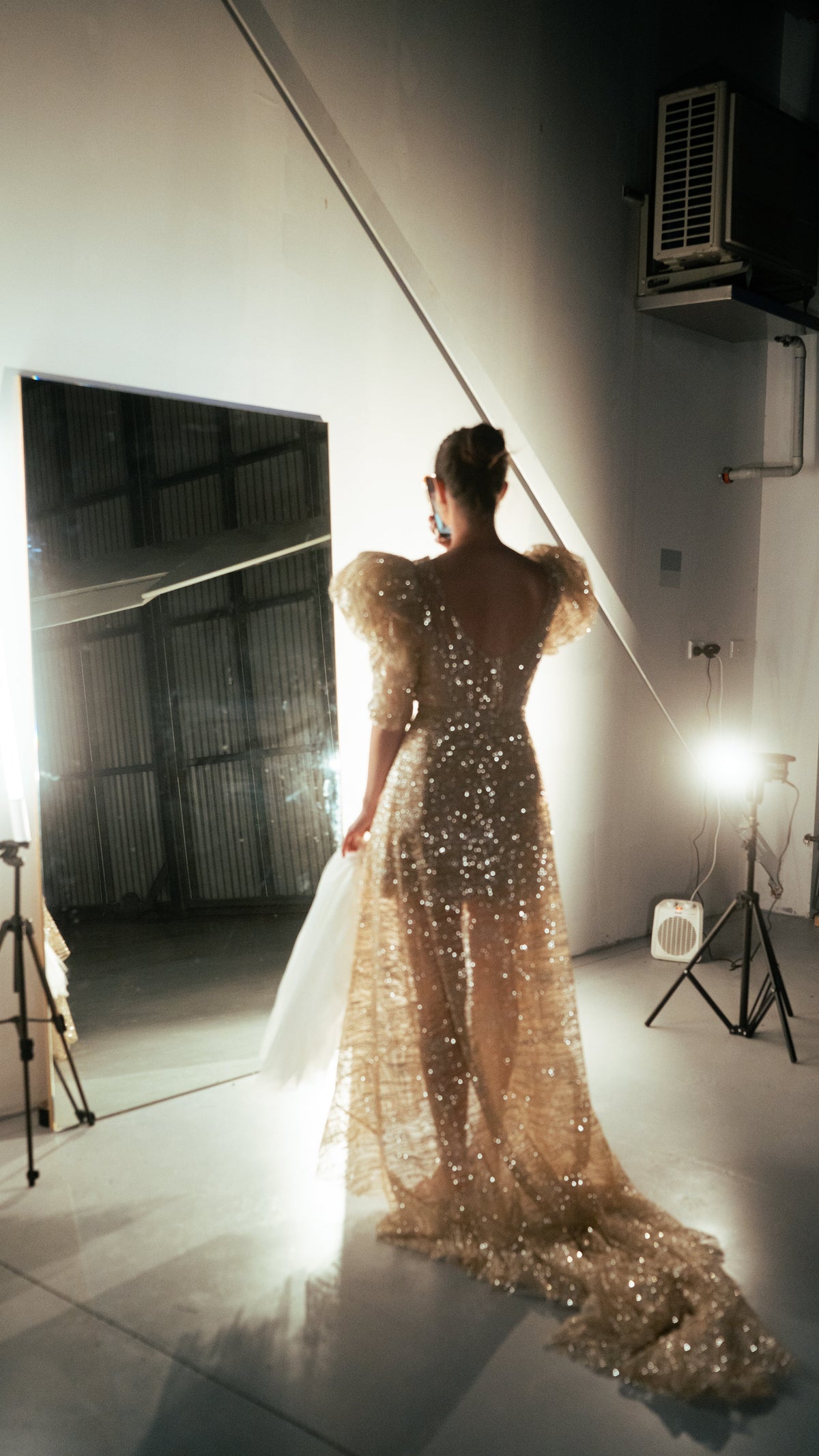Back view of a champagne sparkle wedding dress with puff sleeves and a sheer floor-length overskirt trailing in a lit studio, Soleil by Ephemeris