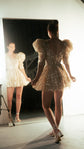 Model admiring a short champagne wedding dress in a studio mirror, puff sleeves and full sparkling skirt, Soleil