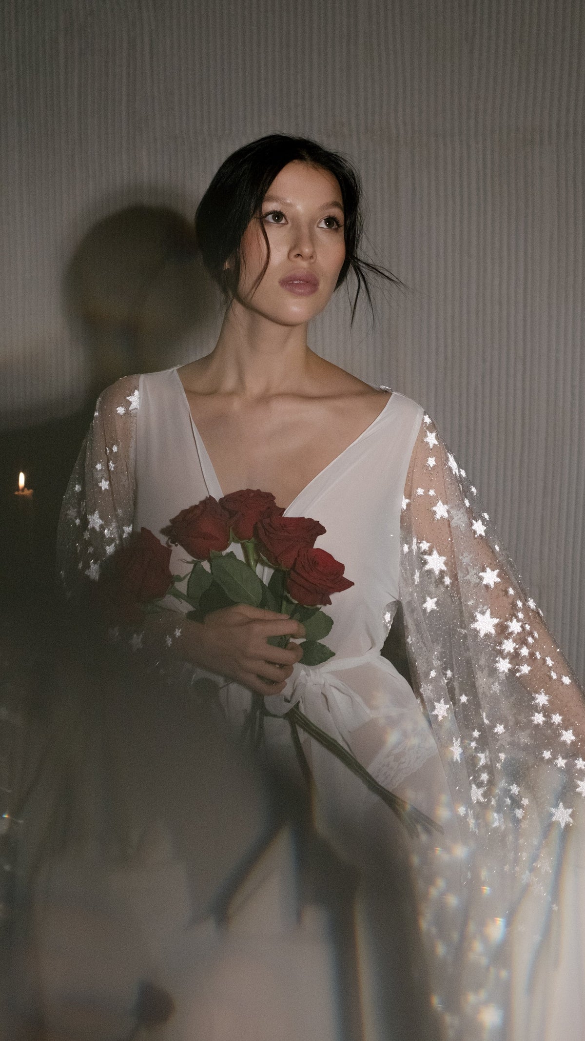 Waist-up portrait in a V-neck bridal robe with star lace sleeves, red roses held to the chest in low candlelight, Stargazer Robe Ivory