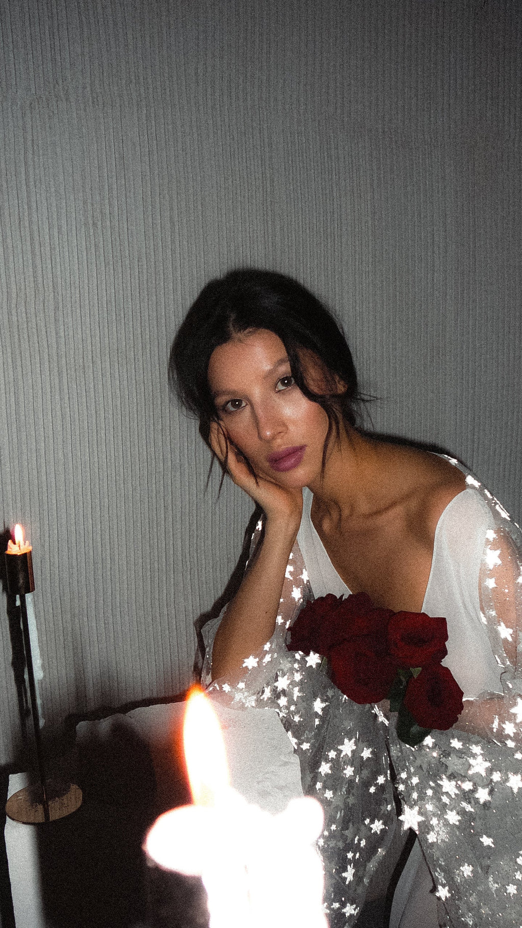 Close portrait in a V-neck chiffon bridal robe, chin in hand behind a candle flame, star lace sleeve and red roses, Stargazer Robe Ivory