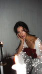 Close portrait in a V-neck chiffon bridal robe, chin in hand behind a candle flame, star lace sleeve and red roses, Stargazer Robe Ivory
