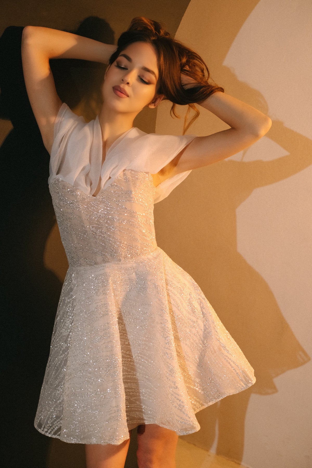 Model with arms raised behind her head in a short sparkle lace wedding dress with organza at the shoulders, warm arch, Starlight