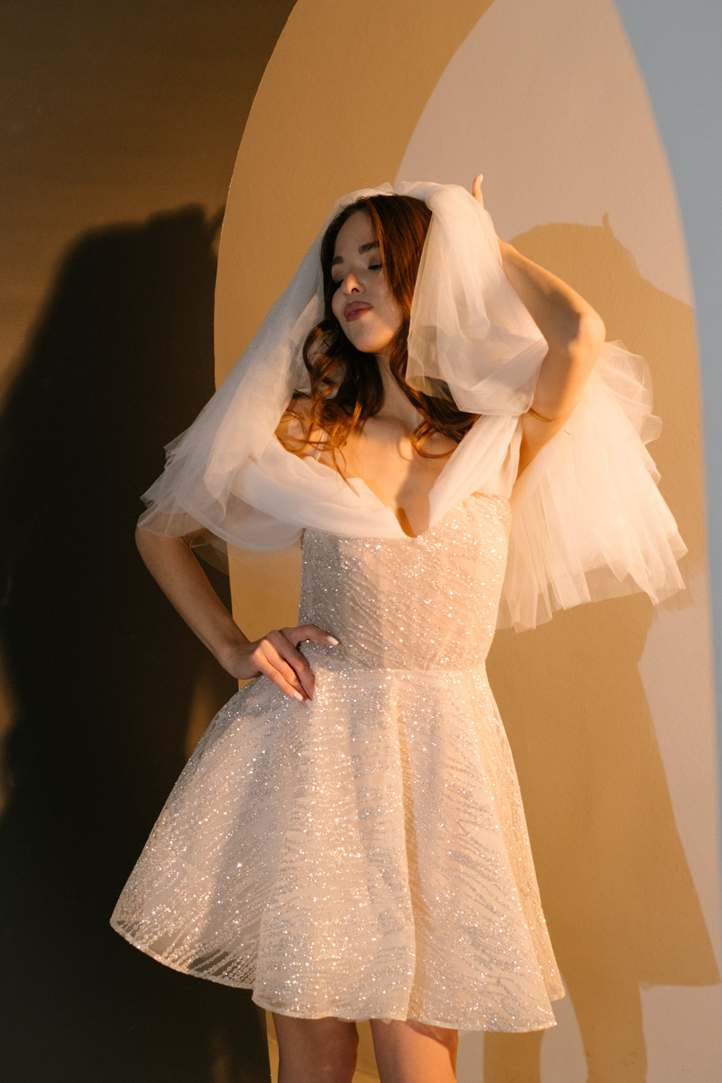 Model holding a short tulle veil over her head in a sparkle lace mini wedding dress, hand on hip, warm arch, Starlight