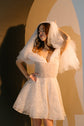 Model holding a short tulle veil over her head in a sparkle lace mini wedding dress, hand on hip, warm arch, Starlight
