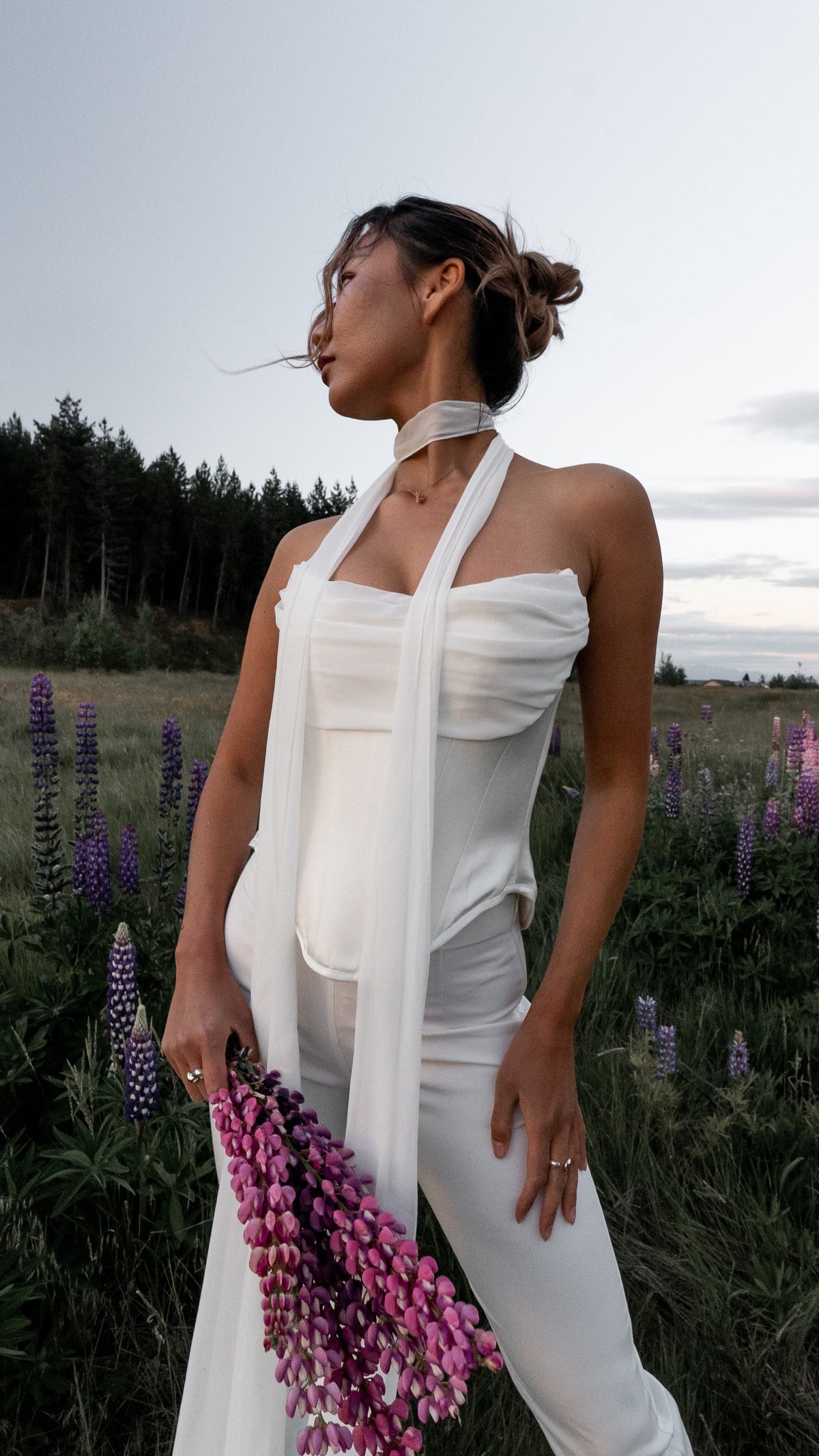 Bride in a draped corset top, long white scarf and ivory bridal trousers, holding pink lupines in a meadow, Sun Flares