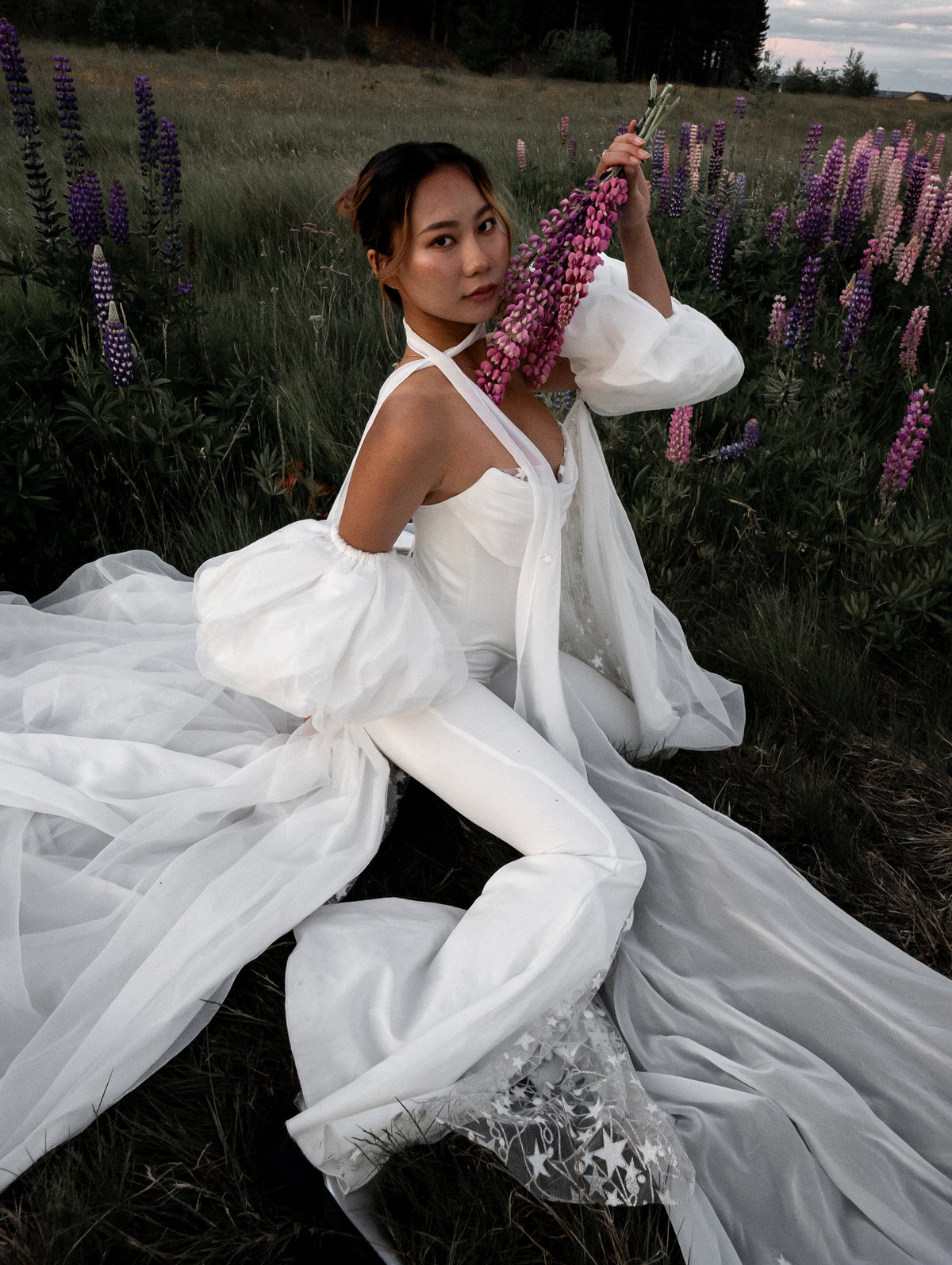 Bride seated in a lupine meadow in bridal trousers with a star lace flare, puff sleeves and a long tulle train, Sun Flares
