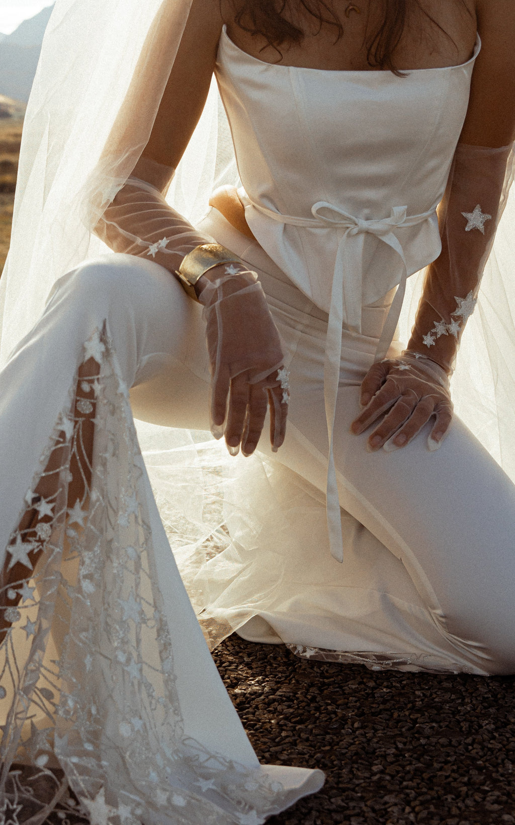 Close-up of star lace in the flared hem of ivory bridal trousers, kneeling bride in a corset top and sheer star gloves, Sun Flares