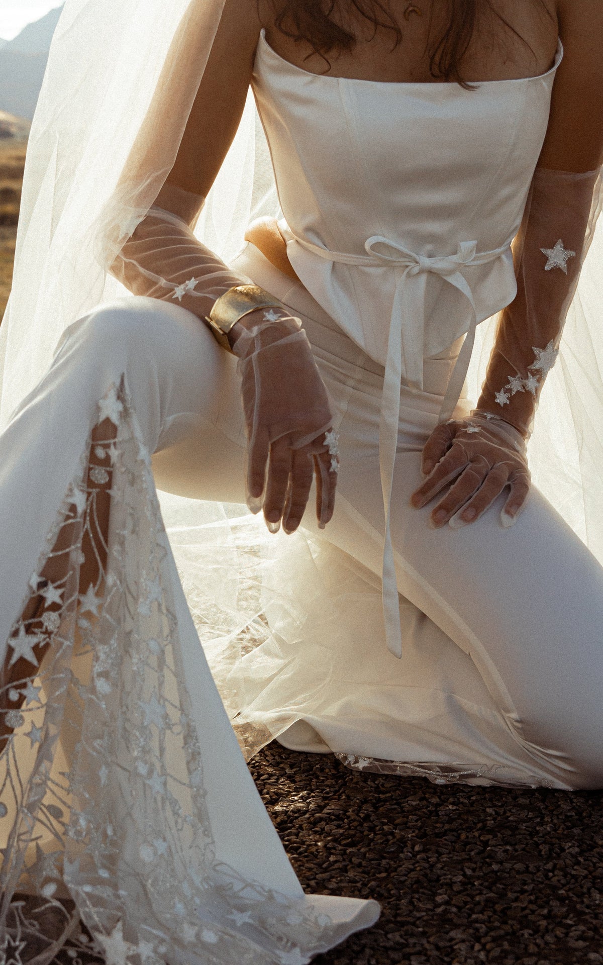 Close-up of star lace in the flared hem of ivory bridal trousers, kneeling bride in a corset top and sheer star gloves, Sun Flares