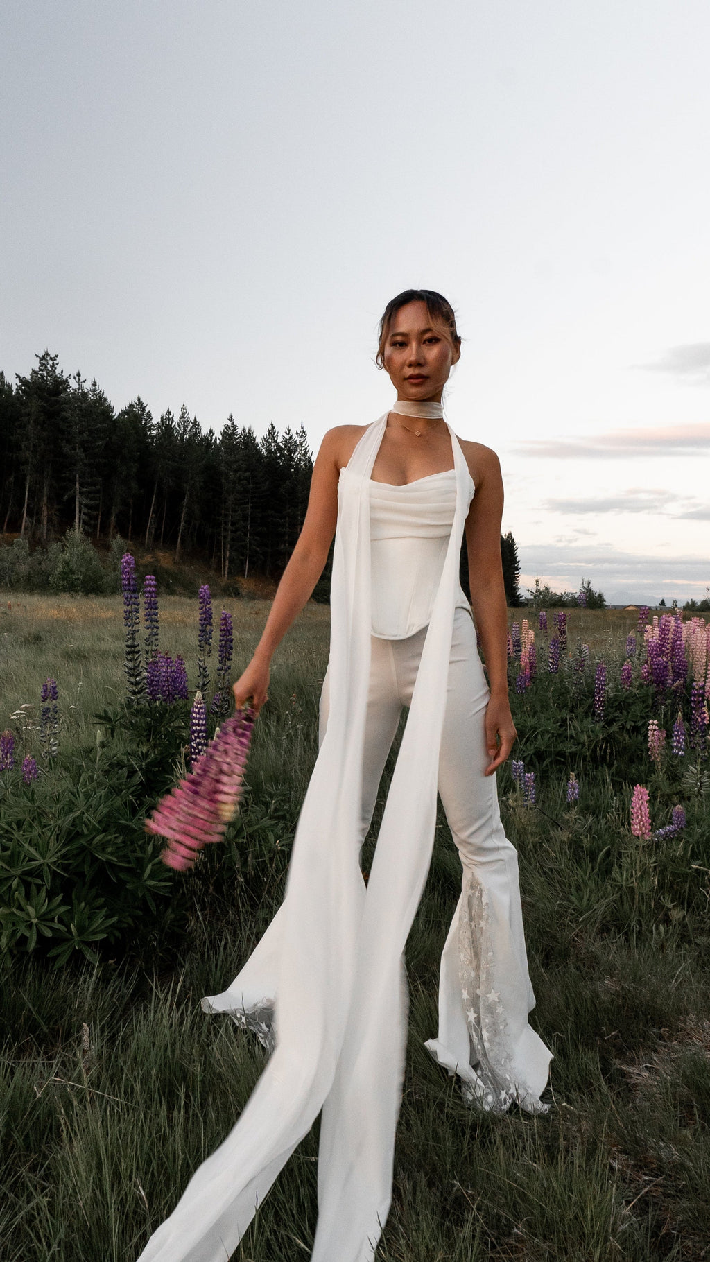 Bride in ivory bridal trousers and a draped corset top with a long scarf in a wildflower field of lupines, star lace flares, Sun Flares