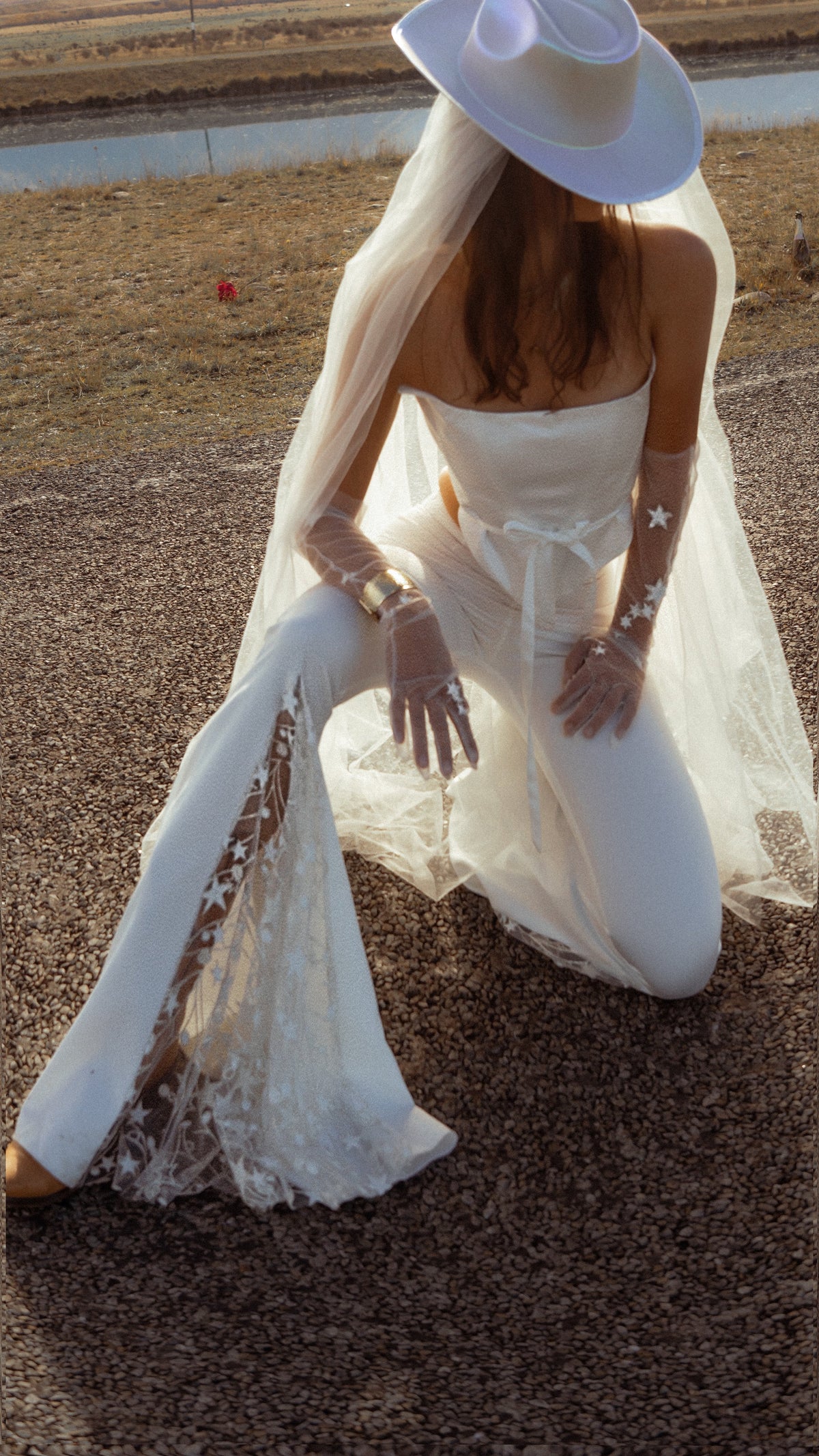 Bride kneeling on a roadside in star lace flared bridal trousers, cowboy hat, tulle veil and sheer star gloves, Sun Flares