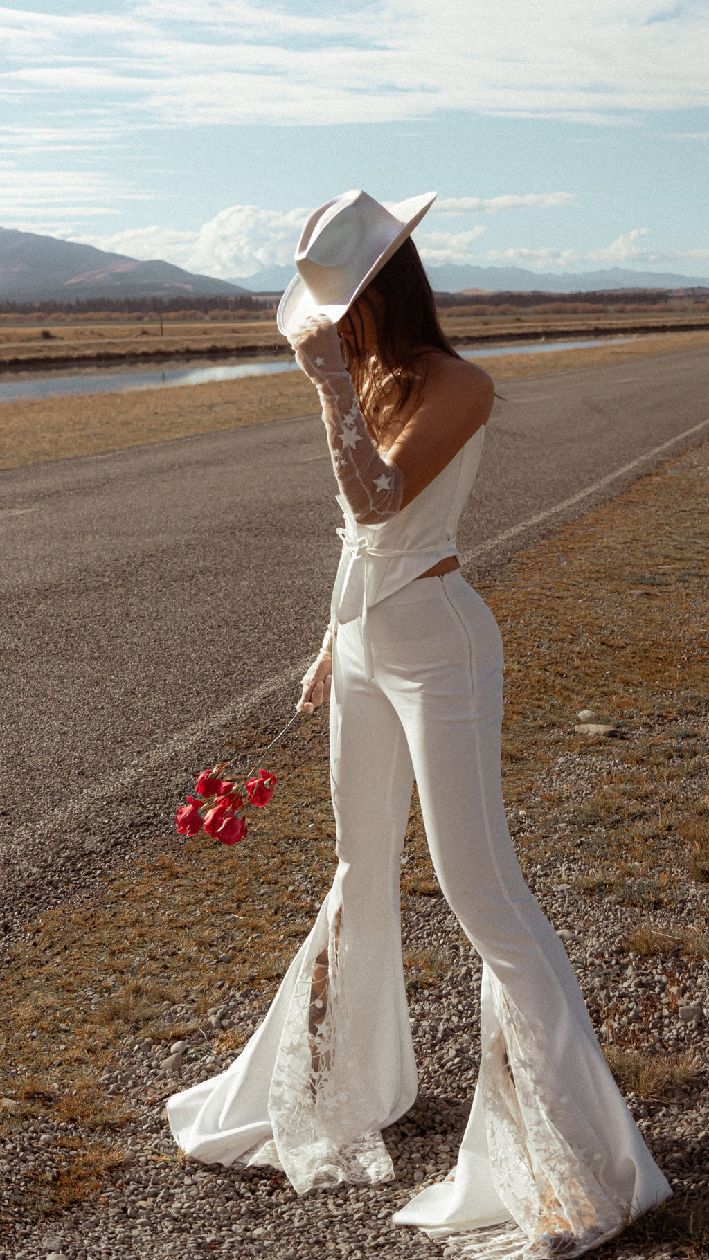 Back view of fitted bridal trousers with a long star lace flare, bride in a cowboy hat holding red roses by a country road, Sun Flares